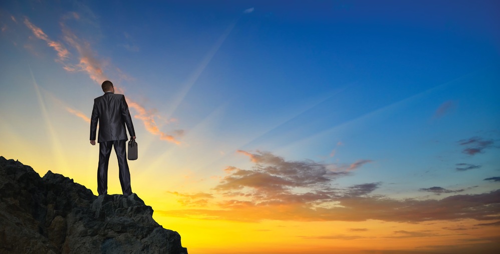 silhouette of a businessman with a laptop in hand is on top of a mountain in the sky with clouds.