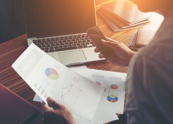 Businessman hands using cell phone with financial report graph at office desk.