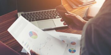 Businessman hands using cell phone with financial report graph at office desk.