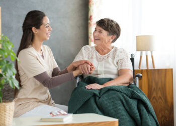 Smiling woman taking care about happy grandmother in wheelchair with green blanket on legs