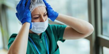 Distraught nurse wearing protective workwear while holding her head in pain at medical clinic.