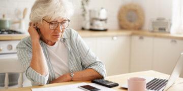 People, age, technology and finances. Depressed unhappy retired woman paying domestic bills online, trying hard to make both ends meet, sitting at kitchen table, surrounded with papers, using gadgets